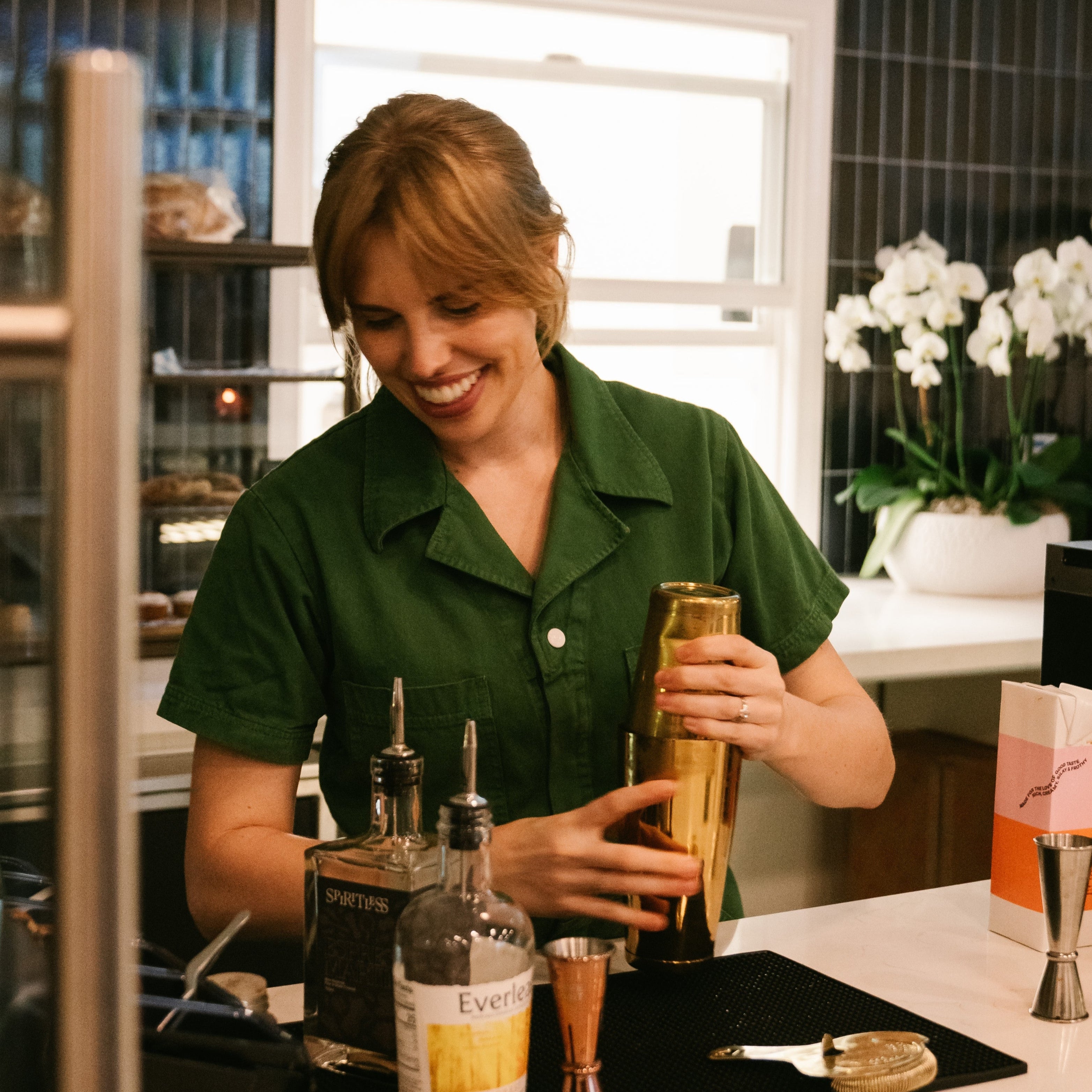 Woman in a green shirt preparing a drink at a bar counter with bottles and a shaker.